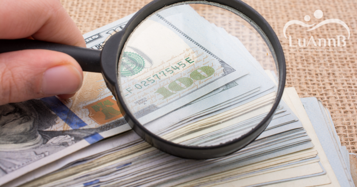 Luanne Blog Post Images-4 Image of a hand holding a magnifying glass over a stack of 100 dollar bills for the post titled Why Passion Alone Won’t Pay the Bills—And What Actually Will by Luann B.