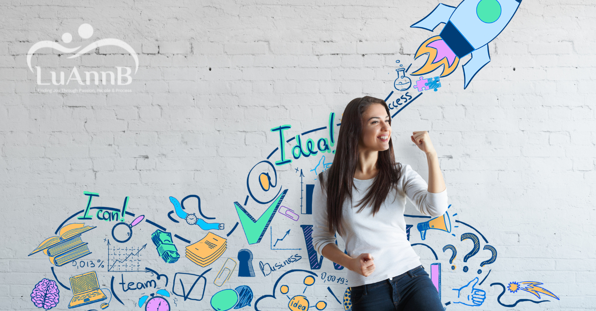 Image of a happy woman with a power pose in front of a wall with motivational business graphics on it. LuAnnB's logo is on the image.